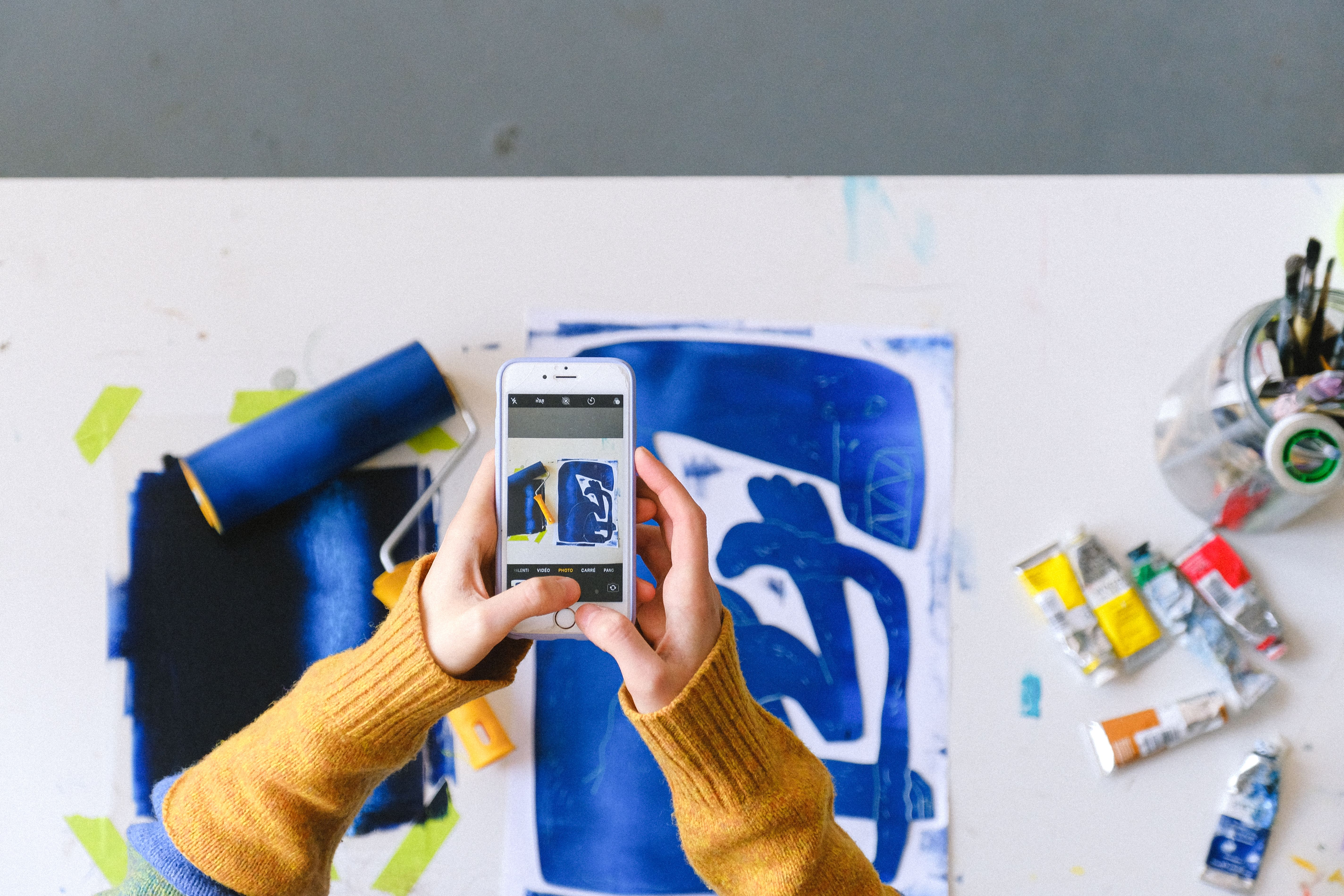 Artist photographing a painted sketch beside paint tubes and brushes
